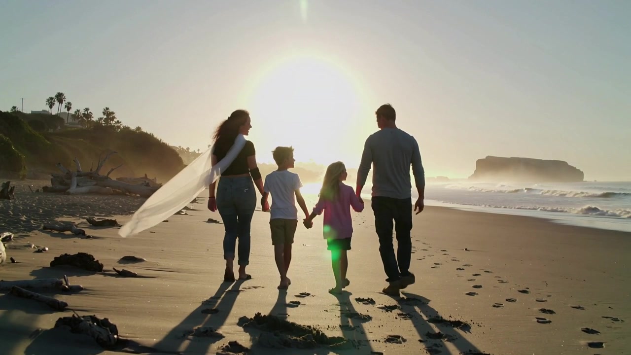 Familia de cuatro caminando siguiendo el sol bajo para llegar a un promontorio en la playa, con una bufanda blanca ondeando detrás