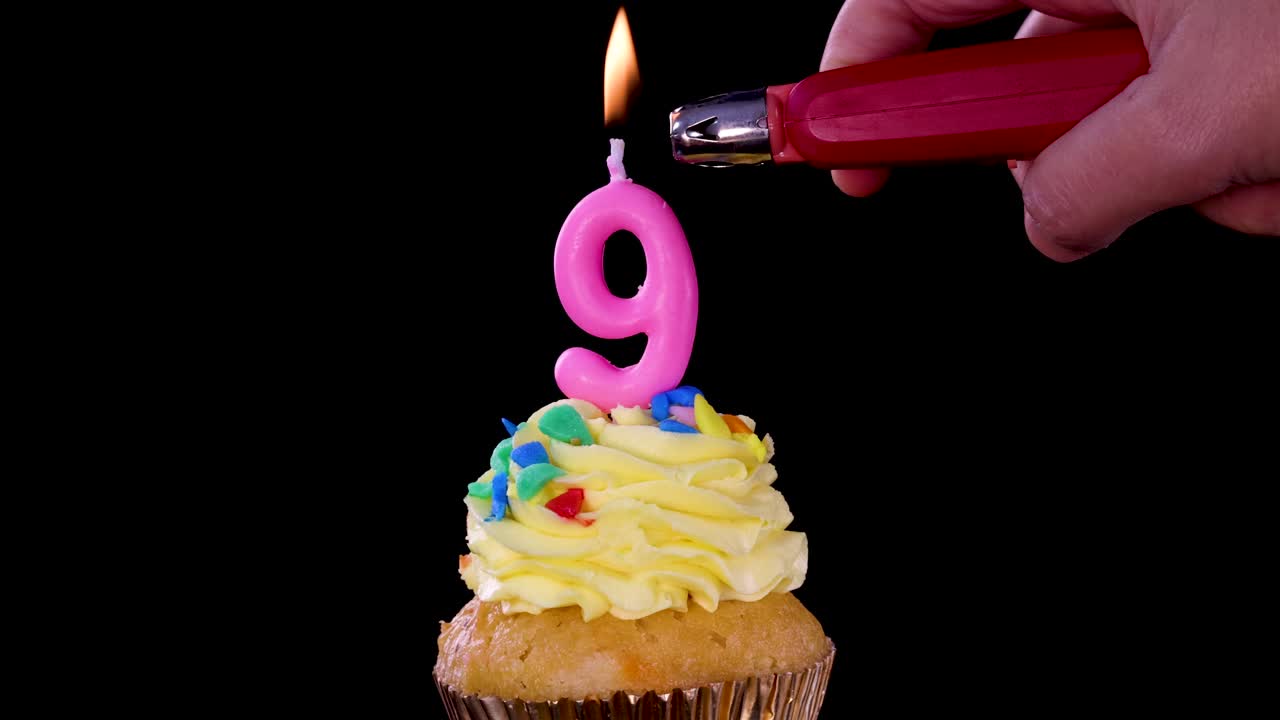 Una mano usa un accendino per accendere una candela rosa a forma di numero nove su un cupcake glassato con zuccherini, su uno sfondo nero con illuminazione stabile.