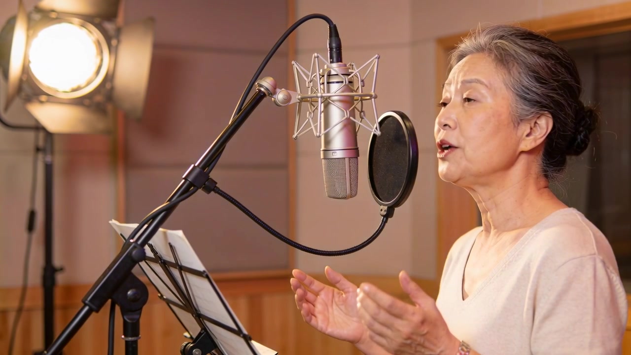 Anziana donna asiatica canta leggendo uno spartito, chinata al microfono in studio per la registrazione delle voci, con spazio per il testo.