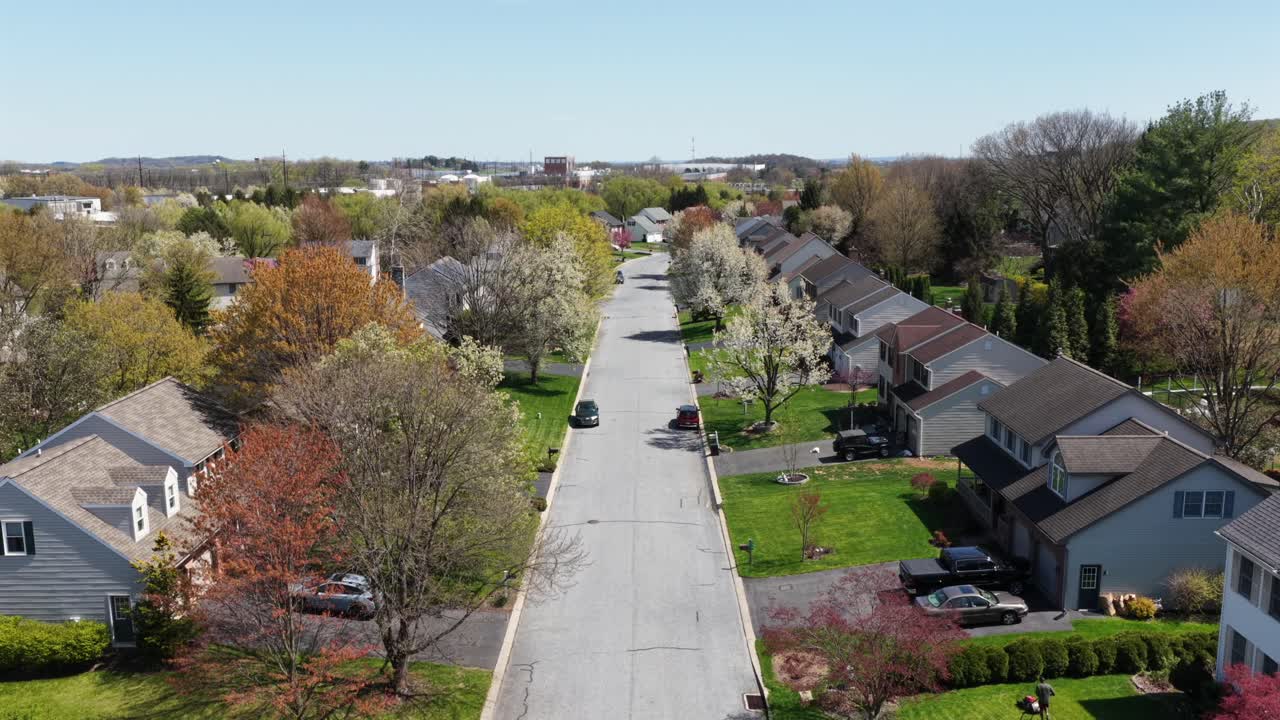 Elevação aérea sobre um bairro suburbano de luxo na Pensilvânia na primavera, com árvores floridas, gramados bem cuidados, casas isoladas e morador cortando a grama em um dia ensolarado. Plano geral