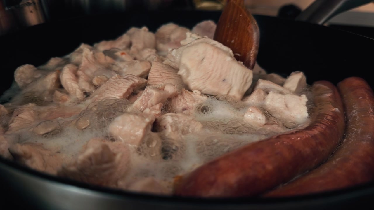 Primo piano di carne di pollo a fette e salsicce che cuociono in una padella calda con vapore e succhi che ribollono. Preparazione di un pasto fatto in casa in cucina durante il processo di cottura