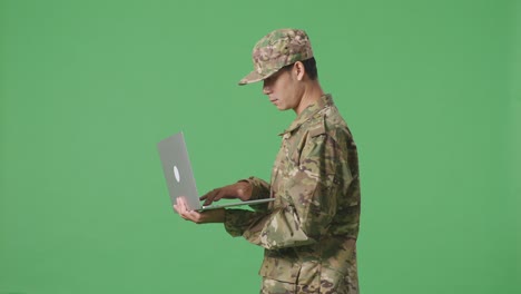 vista lateral de un soldado asiático usando una computadora portátil mientras camina en el estudio de fondo de la pantalla verde