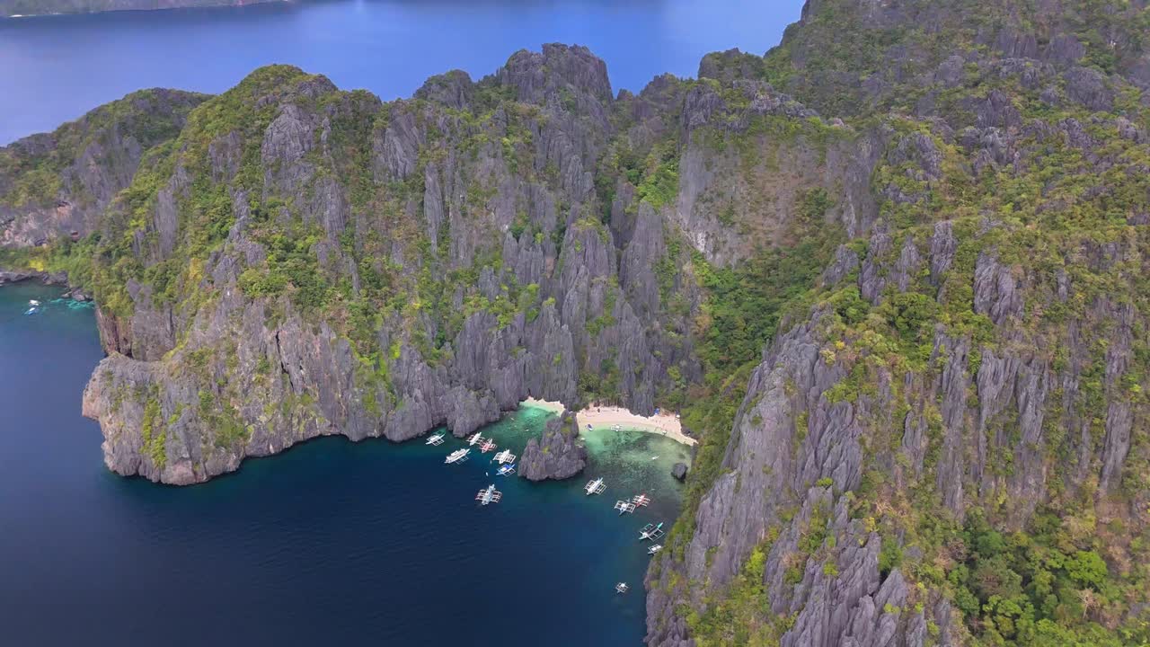 Towering limestone cliffs rise above the turquoise waters and white sand of Jiji's Beach in Secret Lagoon, Miniloc Island, Philippines, with traditional boats anchored in the calm blue bay