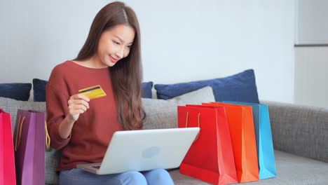 mujer bonita asiática comprando cosas en línea con tarjeta de crédito y portátil con un montón de bolsas de compras en el sofá