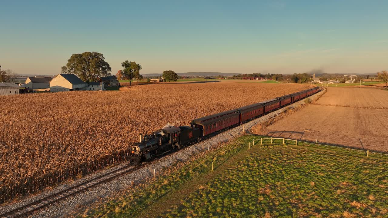 A vintage steam train moves through expansive cornfields, surrounded by trees and distant farms as the sun sets, casting a warm glow over the countryside.