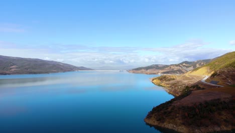 Ruhiges-Wasser-Des-Sees,-Das-Den-Blauen-Himmel-Und-Statische-Wolken-über-Dem-Von-Bergen-Umgebenen-Staudamm-Reflektiert