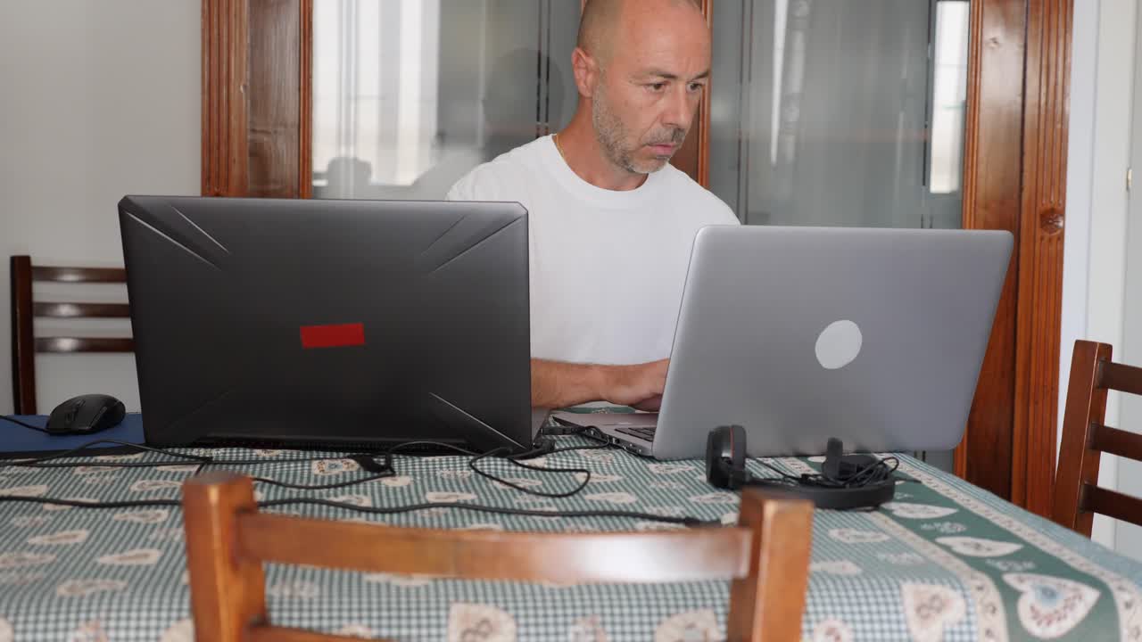hombre calvo de mediana edad que trabaja con dos computadoras portátiles en casa