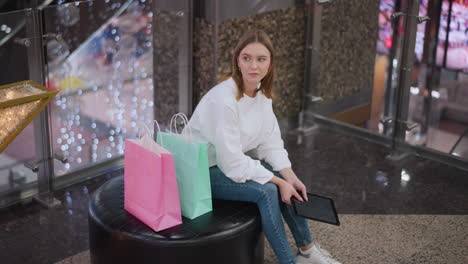 young woman seated in mall holding tablet, looking contemplative with a warm smile, surrounded by shopping bags and festive decorations, the background features glowing lights