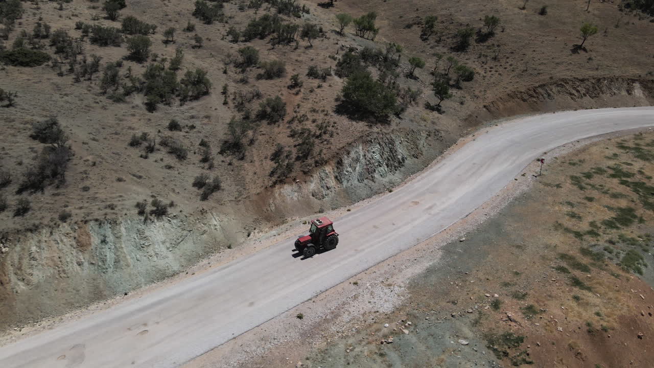 Drone che riprende un trattore rosso che avanza lentamente su una strada tra campi e montagne a Karaman, Turchia, in una soleggiata giornata estiva.
