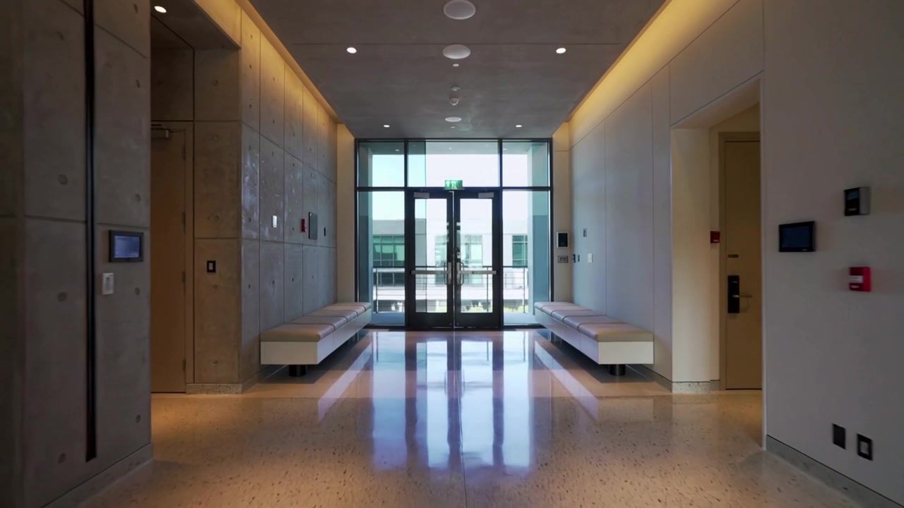 La telecamera si allontana, rivelando doppie porte d'ingresso in vetro in un corridoio di un edificio moderno, con panchine.