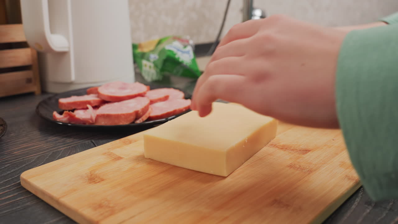 close up of woman in green sleeve holding yellow butter block while removing plastic wrapper over wooden board in kitchen, with sliced sausage and white electric kettle visible in background