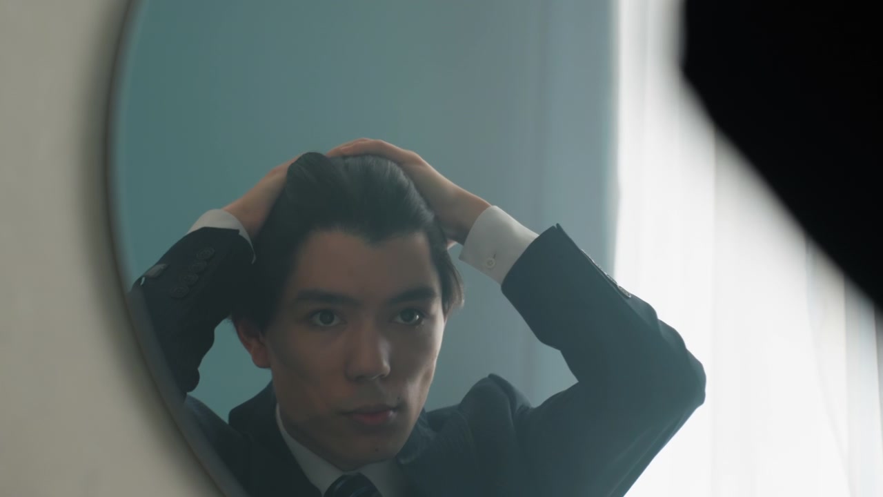 Asian man in suit checking reflection in round mirror adjusting hair and tie with soft window backlight, practicing expressions and composure, scenes suggest interview rehearsal, actor warmup