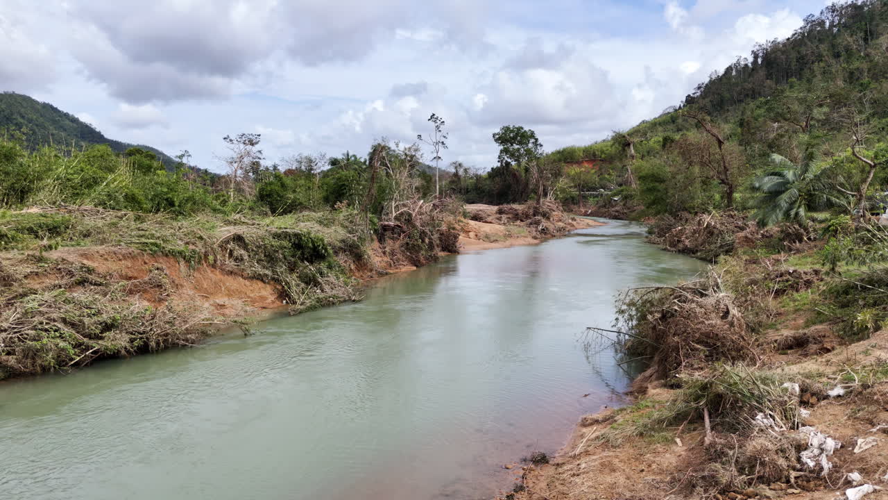 Plano de drone de baixa altitude seguindo o Rio Cataban, mostrando vegetação danificada e erosão do solo após as inundações do Tufão Kalmaegi.