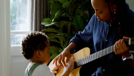 Vista-Lateral-De-Una-Joven-Madre-Negra-Y-Un-Hijo-Pequeño-Tocando-La-Guitarra-En-El-Salón-De-Una-Cómoda-Casa-4k