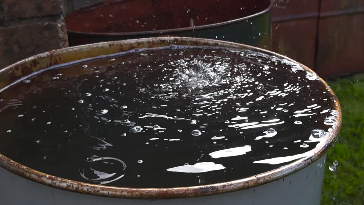 Water dripping and falling into a metal barrel after a rainy day in slow motion. The imapct creates water ripples and small waves that bounce and water is flowing out from the barrel. Shallow depth