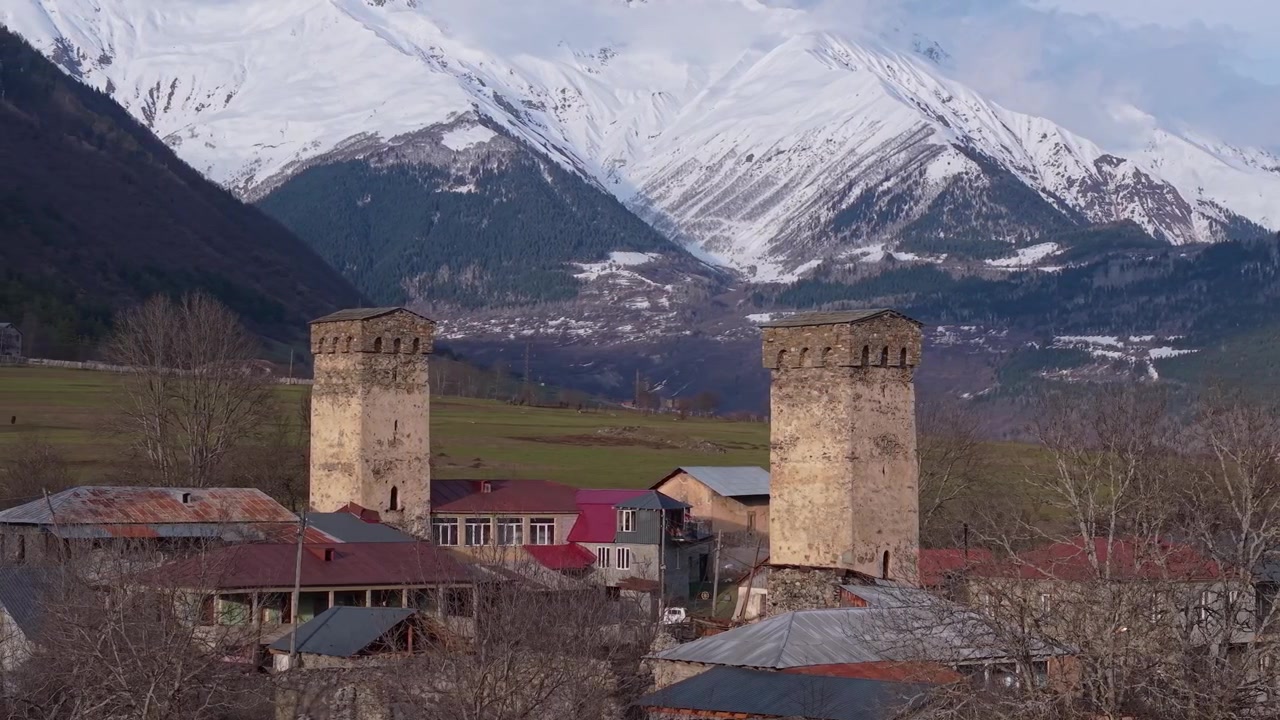 Vista aérea de casas tradicionais na região de Svaneti, na Geórgia, com uma icônica torre Svan, vales exuberantes e as montanhas do Cáucaso cobertas de neve criando um cenário espetacular.
