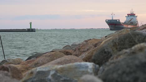 large gray cargo ship entering the port of liepaja in calm sunny summer evening, stone pier in foreground, waves splashing, distant medium low angle shot