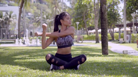 Asian-sportive-woman-sitting-with-crossed-legs-and-stretching-arms-in-park