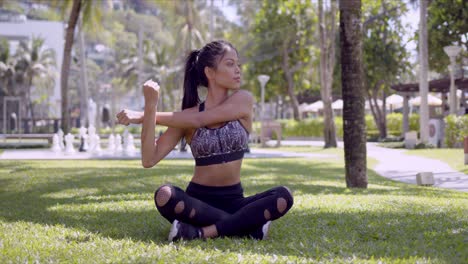 mujer deportiva asiática sentada con las piernas cruzadas y estirando los brazos en el parque