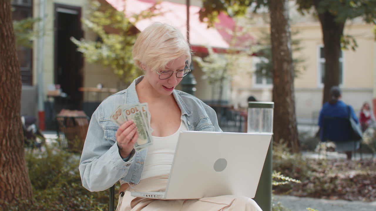Happy woman holding cash while working on laptop outdoors