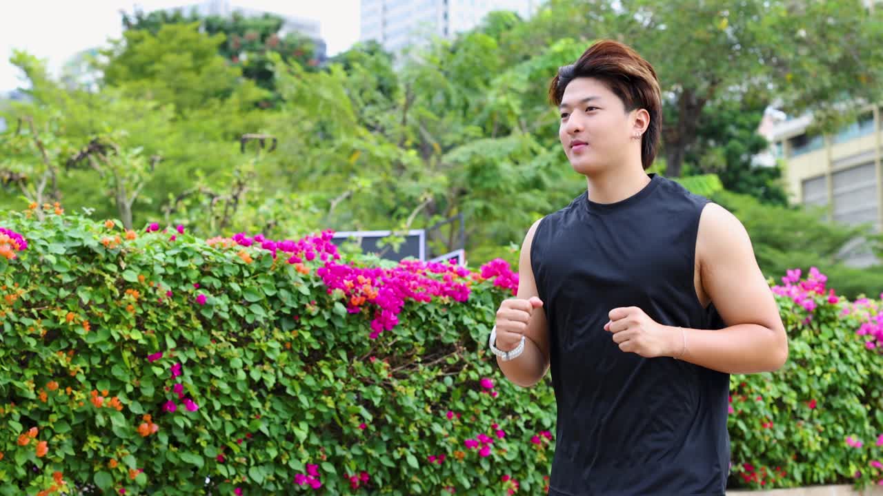 ชายเอเชียรูปร่างดีวิ่งเหยาะๆ ไปตามแปลงดอกไม้ในสวนสาธารณะในเมืองที่สดใส ภาพมุมราบตามติด