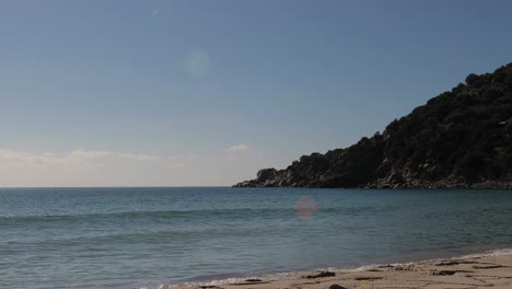 Sandy-beach-scene-with-calm,-tranquil-sea-waves-sparkling-in-bright-tropical-sunshine-with-a-rocky-mountain-in-background