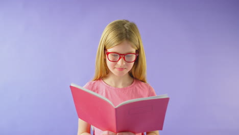 foto de estudio de una niña con gafas estudiando el libro de ejercicios escolar contra un fondo púrpura