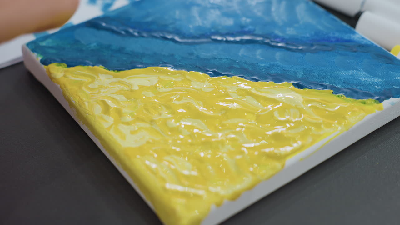 캔버스 가장자리에서 두꺼운 노란색과 파란색 페인트를 다듬고 모양을 만드는 클로즈업된 흰색 여성의 팔뚝, 촉각적인 팔레트 나이프 텍스처, 스튜디오 테이블 및 툴이 보이고 실험적인 분위기