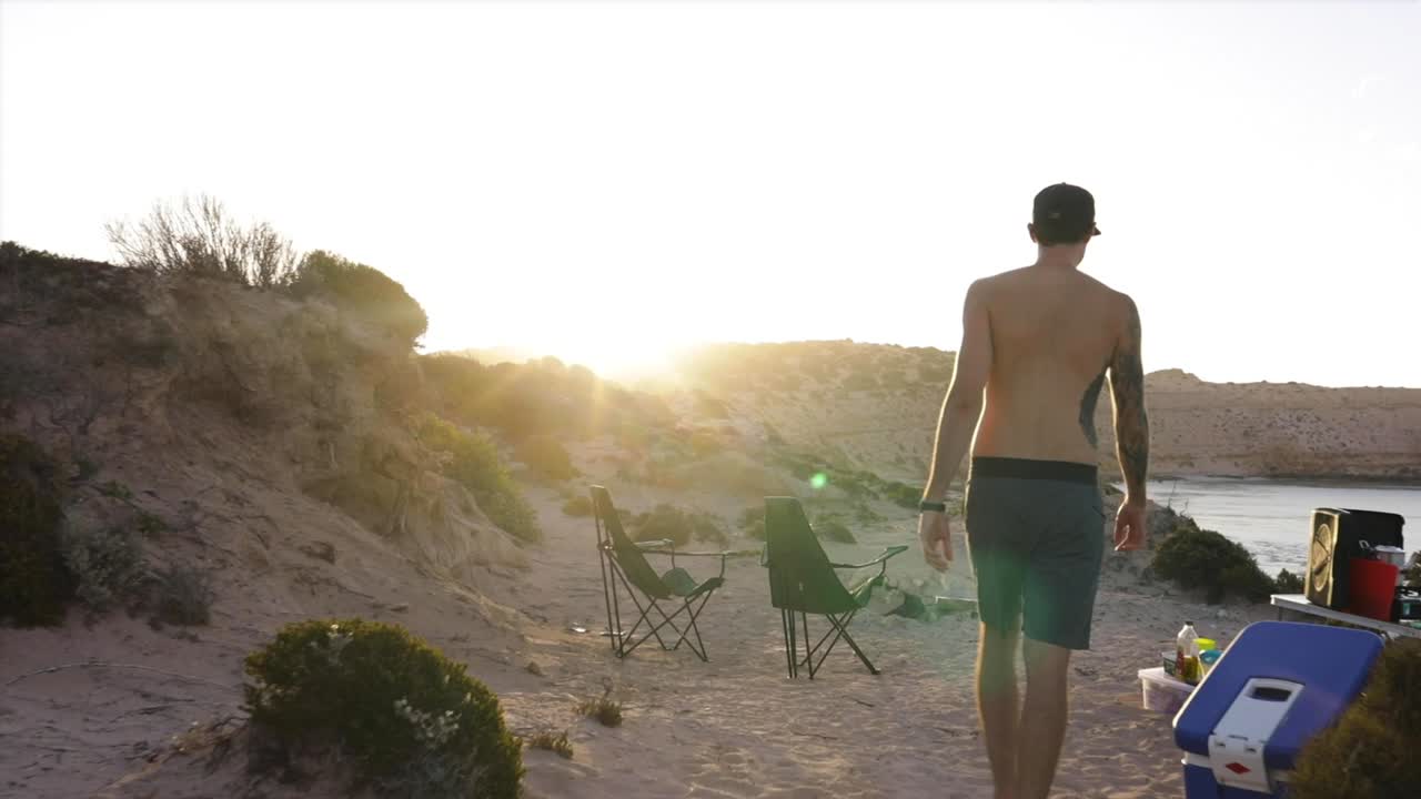 카메라가 아름다운 바다 전망을 바라보는 동안 한 청년이 해가 질 때 절벽 꼭대기 캠프장을 걸어가 불 옆에 앉는다