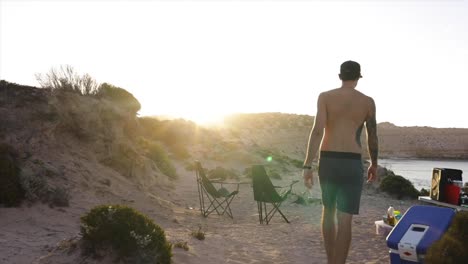 un joven camina por un campamento en lo alto de un acantilado al atardecer, se sienta junto al fuego mientras la cámara descansa sobre hermosas vistas al océano