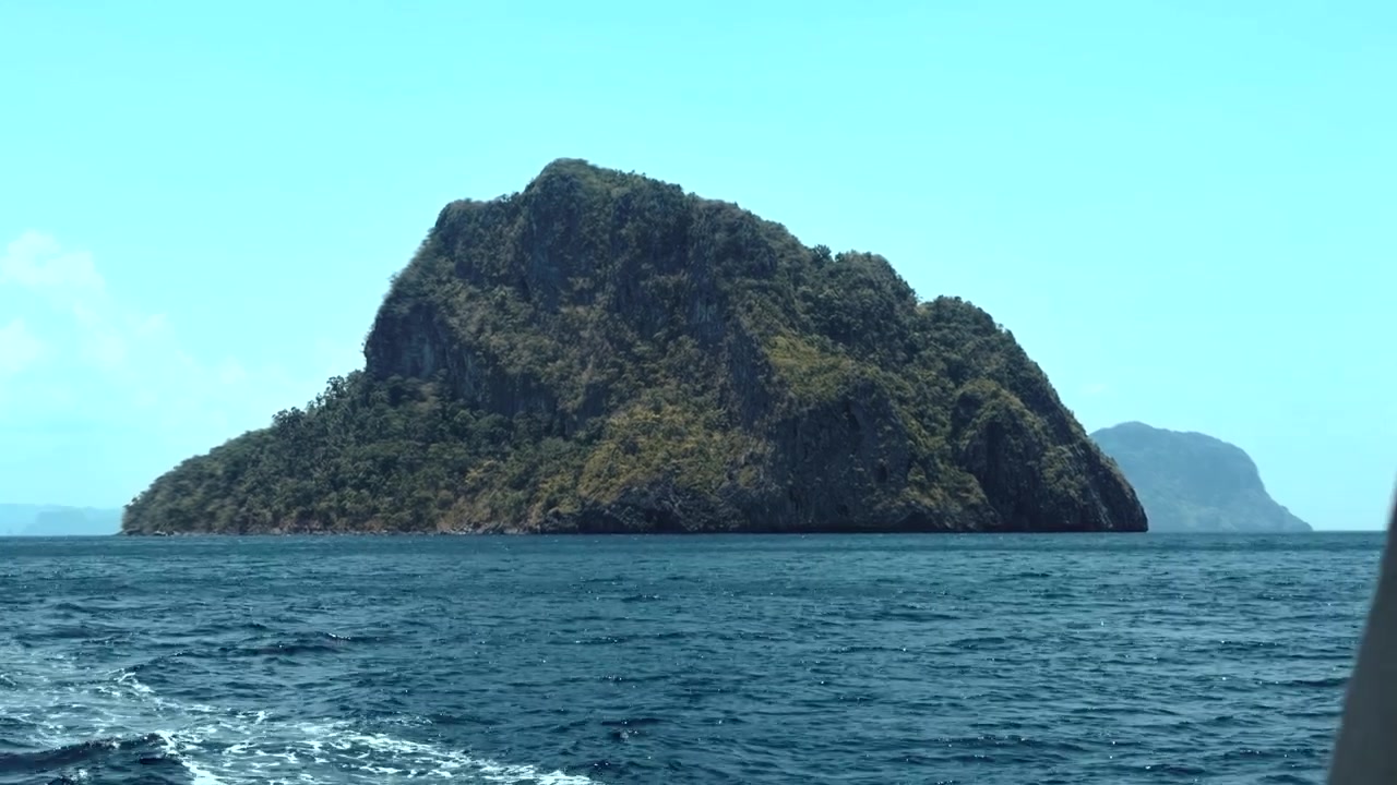 Plynulý boční přelet přes vápencový ostrov pokrytý bujnou vegetací, vystupující z klidných modrých oceánských vod. · 4K DCI 25fps