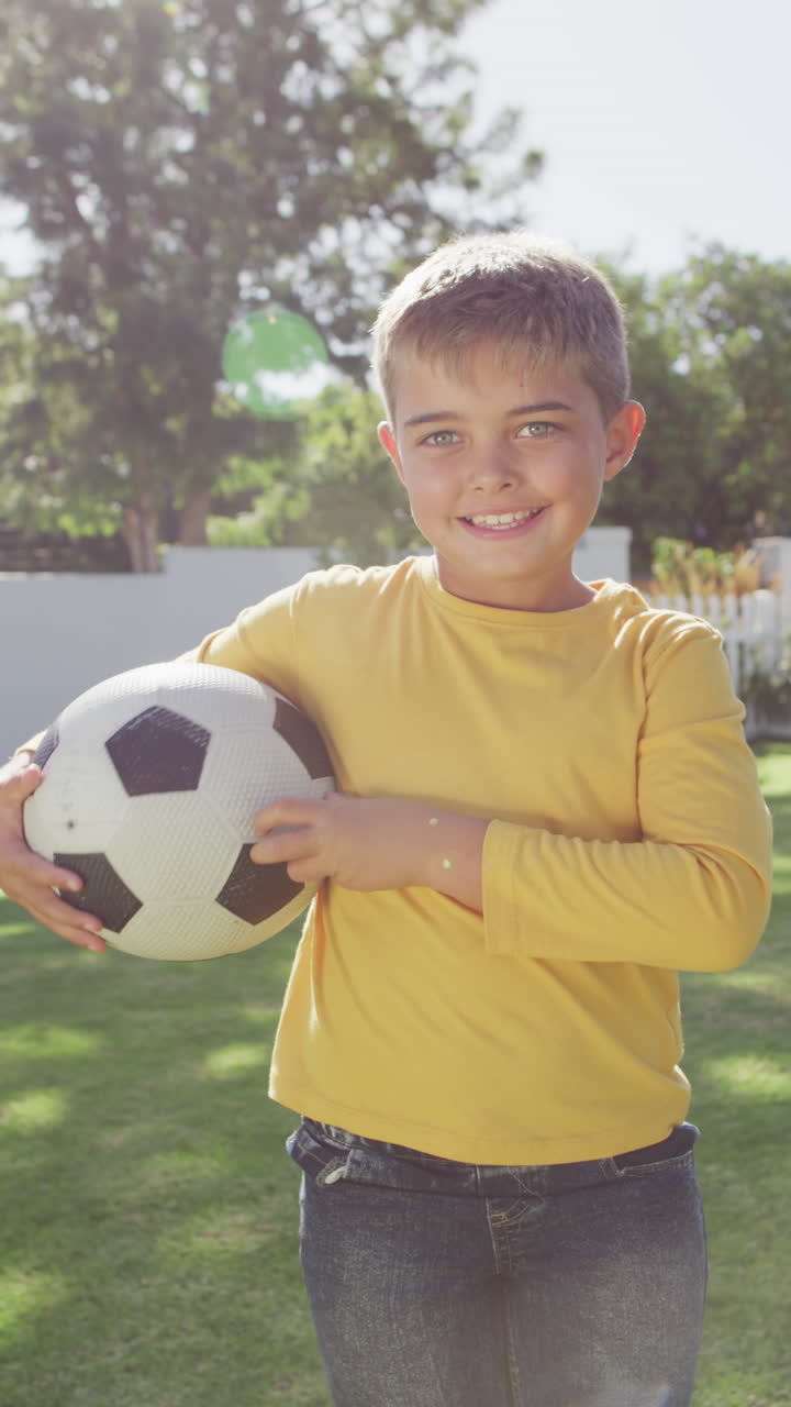 Vertical video of portrait of happy caucasian boy holding football in garden, slow motion