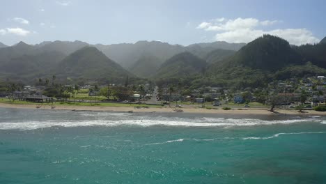 Aerial-shot-over-the-turquoise-waters-of-the-pacific-ocean-going-toward-the-community-of-Hauula-Hawaii-with-the-dense-mountains-in-the-background