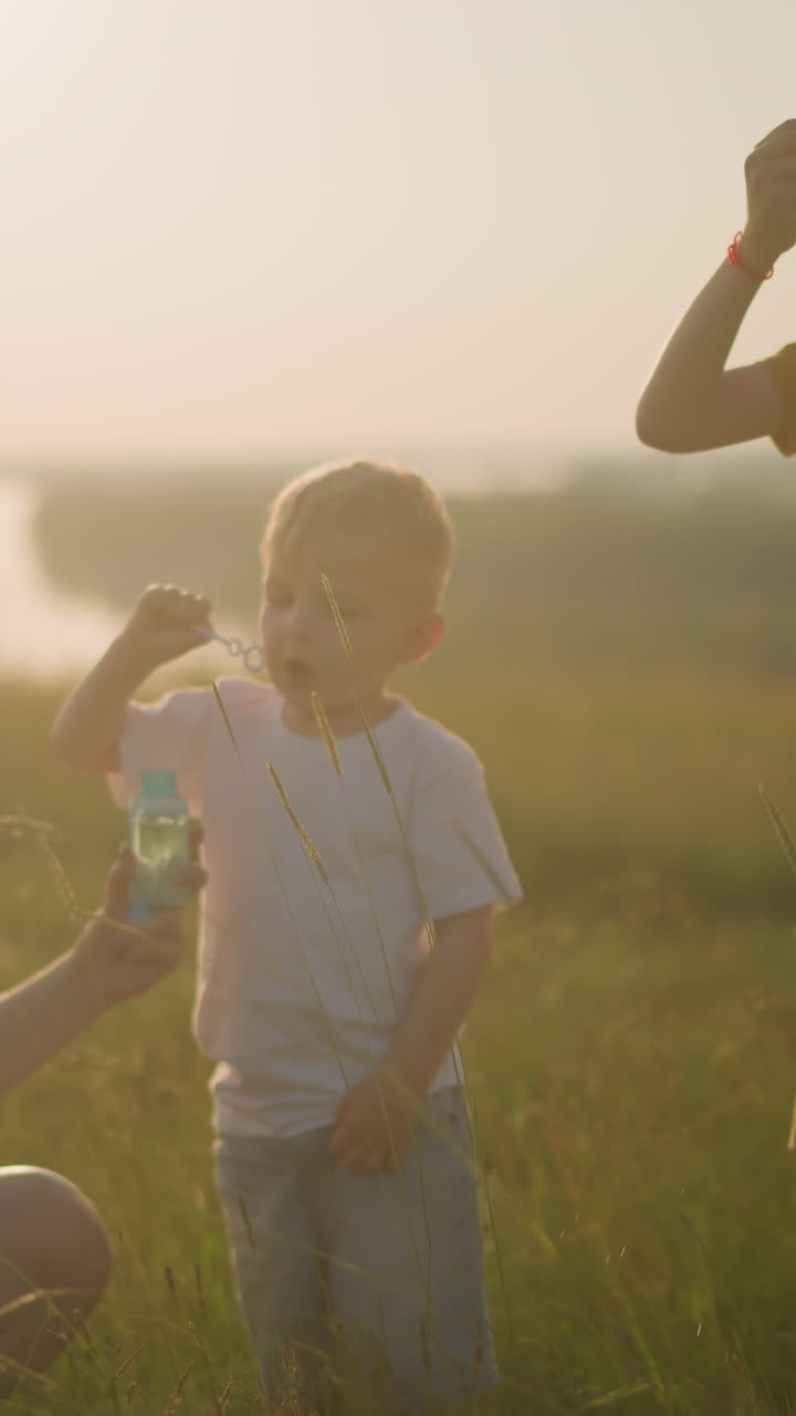 радостная женщина в синем платье коленичит в травяном поле на закате, держа в руках бутылку с пузырьками, в то время как ее младший сын погружает палочку и дует пузырьями. ее старший сын, одетый в желтый наряд, также дует пузыри