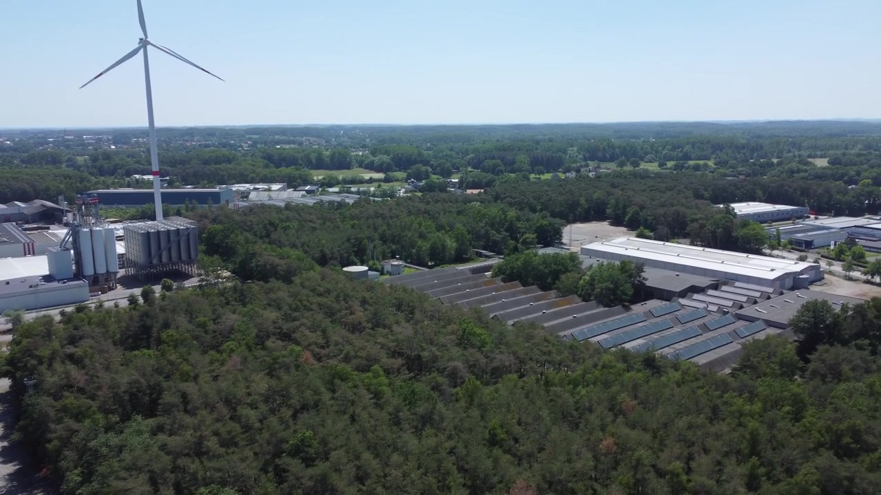 Drone footage starting above a highway beside warehouses and storage silos, flying over dense trees to reveal another industrial site with additional warehouses.