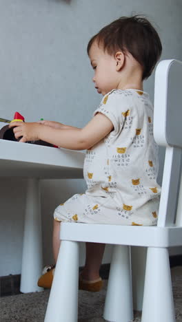 toddler boy wearing white clothes sits at the desk. small kid plays bright toys. low angle view. vertical video