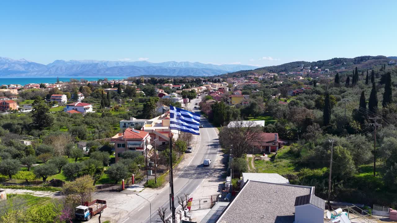 Yunanistan'ın Kuzey Korfu bölgesindeki Acharavi'nin havadan geniş çekim görünümü, dalgalanan Yunan bayrağı, sahil kasabası, yol trafiği, İyon Denizi ve dağları.
