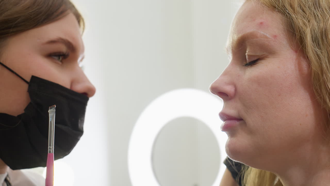 Beautician wearing black mask and gloves prepares to clean excess eyebrow product from client face using cotton pad while holding precision brush in other hand, illuminated by ring light in background