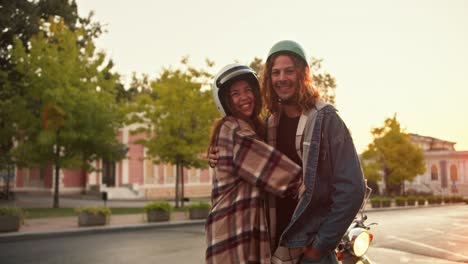 retrato de un chico feliz con el cabello rizado en un casco y una camisa de vaqueros se encuentra al lado y abraza a su novia morena con el pelo rizado en una camisa a cuadros y un casco blanco cerca de su motocicleta en una calle ancha