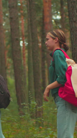 joven con mochila roja ayuda a su amiga a ajustar su bolsa negra en un entorno pacífico del bosque, la amiga con bufanda azul se da la vuelta y sonríe cálidamente antes de caminar juntos
