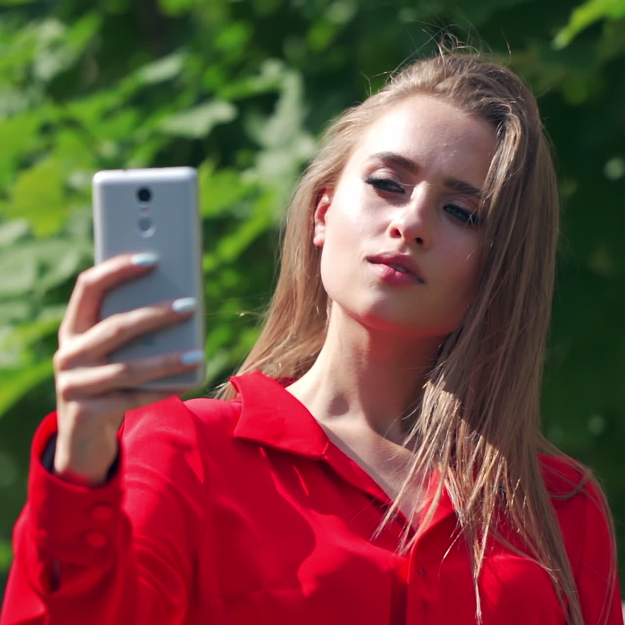 Beautiful girl doing selfie outdoors. Attractive female holding mobile phone in her hand and photographing herself near the green tree. Close-up.