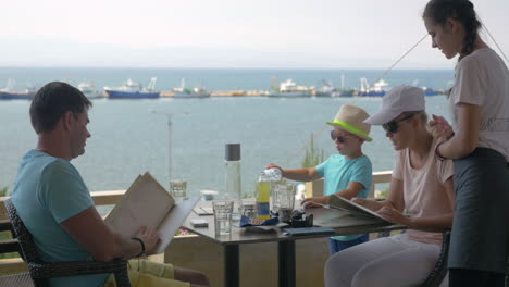 En-La-Costa-Del-Mar-De-La-Ciudad-De-Perea,-Grecia,-Una-Joven-Familia-Se-Sienta-En-Un-Café.