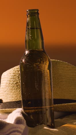 Vertical-Video-Summer-Holiday-Concept-Of-Beer-Bottle-On-Beach-Towel-With-Sun-Hat-Against-Sunset-Sky