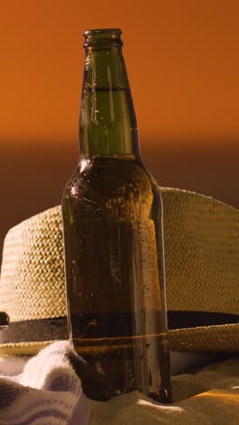 video concepto de vacaciones de verano vertical de botella de cerveza en la toalla de playa con sombrero de sol contra el cielo al atardecer