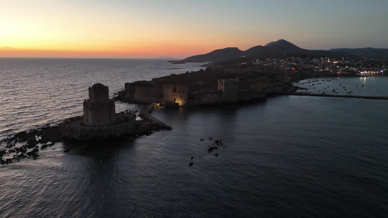 Methoni, Messenia, Peloponneso. Ripresa aerea con panoramica circolare a sinistra intorno alla torre di Bourtzi, dietro si trova il castello di Methoni durante il tramonto. Cielo colorato e mare blu scuro. Luci accese in città.