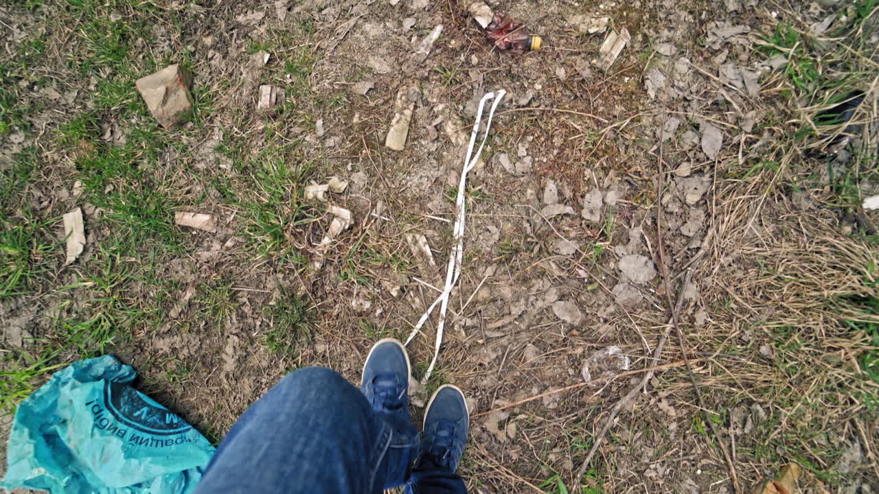 Man's legs in jeans and trainers walking along the dirty place with a lot of rubbish. Plastic and glass bottles and other debris scattered on the ground by people.