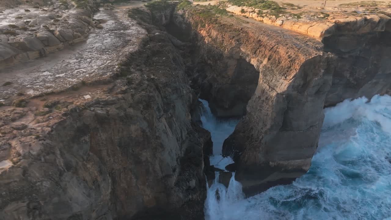 Aerial drone of powerful blue ocean waves hitting rocky sandstone cliffs and a natural stone bridge during sunset, Gozo, Malta, Wied il Mielah, Coastal erosion and climate change, sea level rise