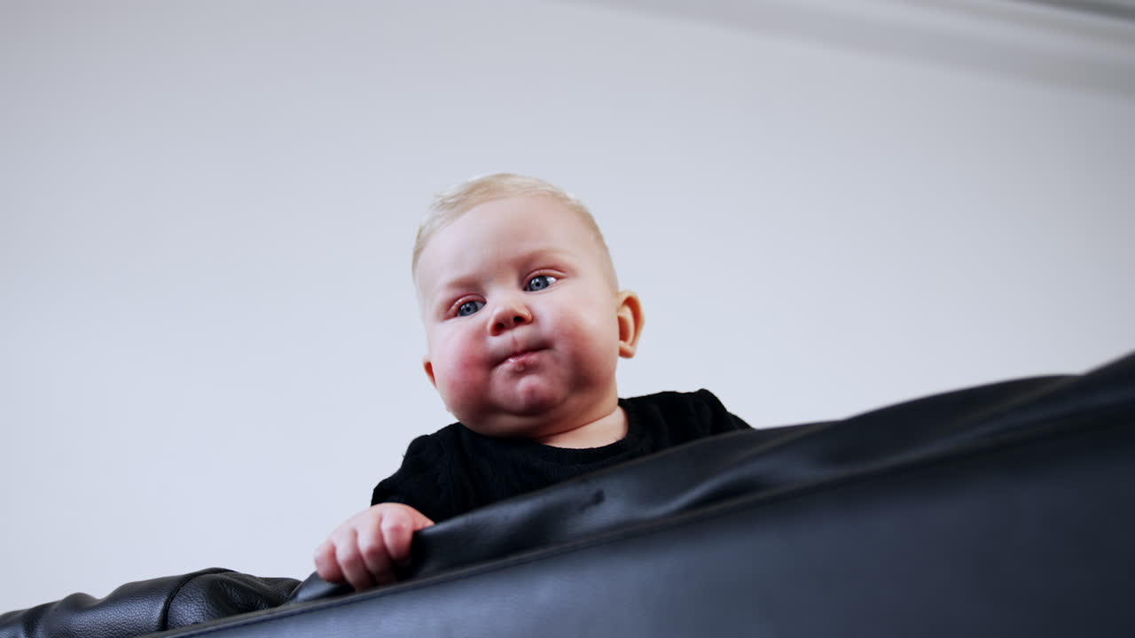 Small blond baby boy with plump cheeks is on the black leather sofa. Low angle view on the child of eight months old.