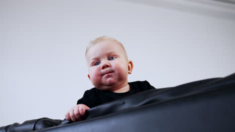 small blond baby boy with plump cheeks is on the black leather sofa. low angle view on the child of eight months old.