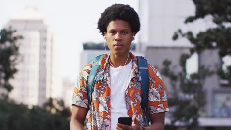 feliz hombre afroamericano en la ciudad, usando auriculares, usando teléfono inteligente y riendo en la calle